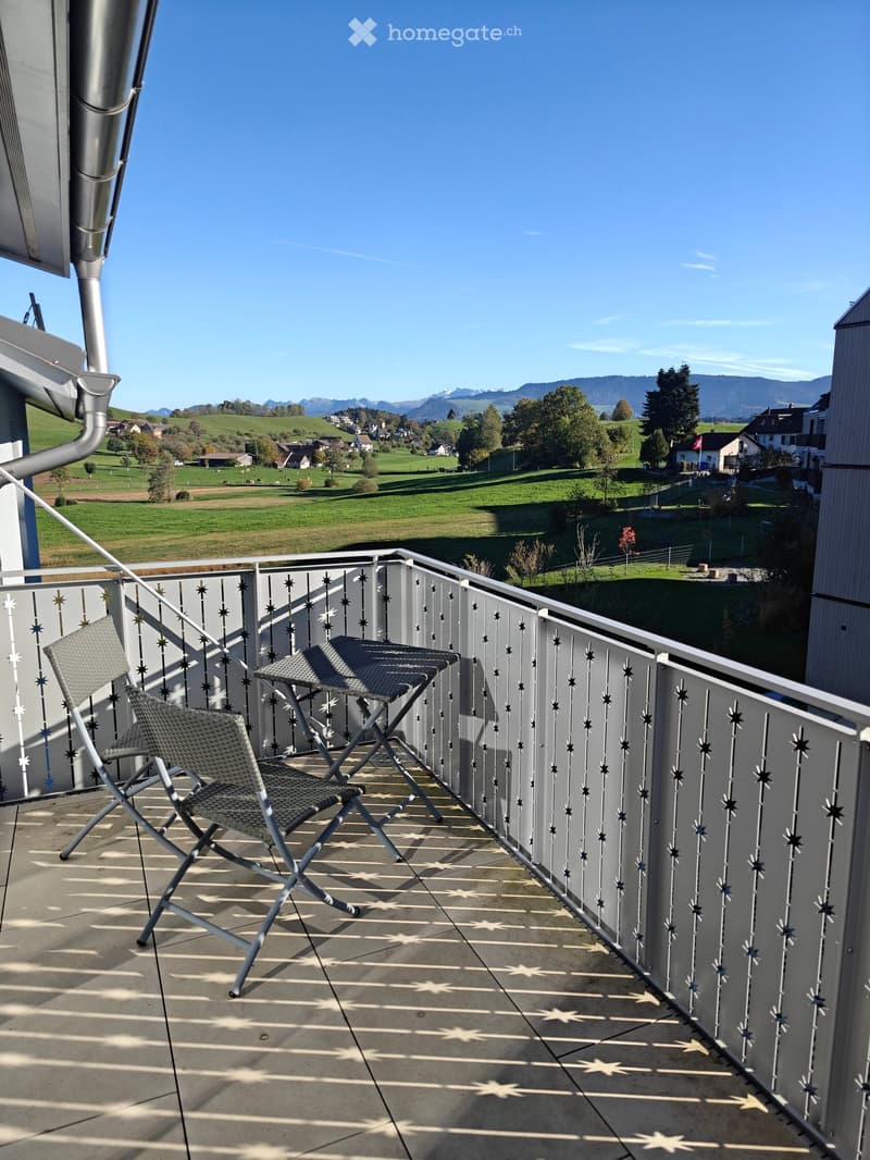 Balcony with a view of green fields and mountains, featuring two folding chairs and a small table.