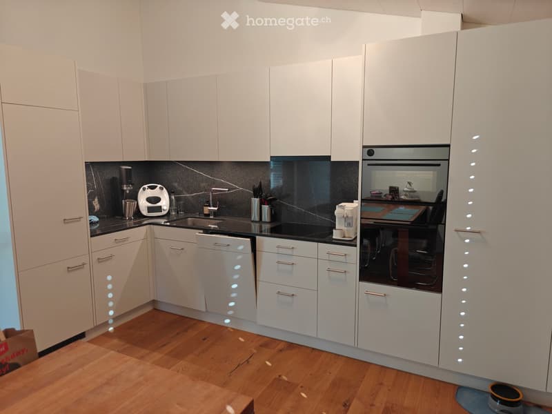 Modern kitchen with white cabinets, black countertops, and stainless steel appliances including an oven, dishwasher, coffee maker, and toaster. The floor is wooden, and the backsplash is dark.