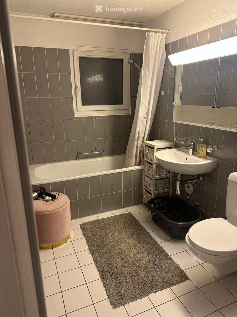 Modern bathroom with a bathtub, sink, toilet, and a small storage unit. Features a grey tiled wall, a window, a pink stool, a grey bathroom rug, and a black shower basket.