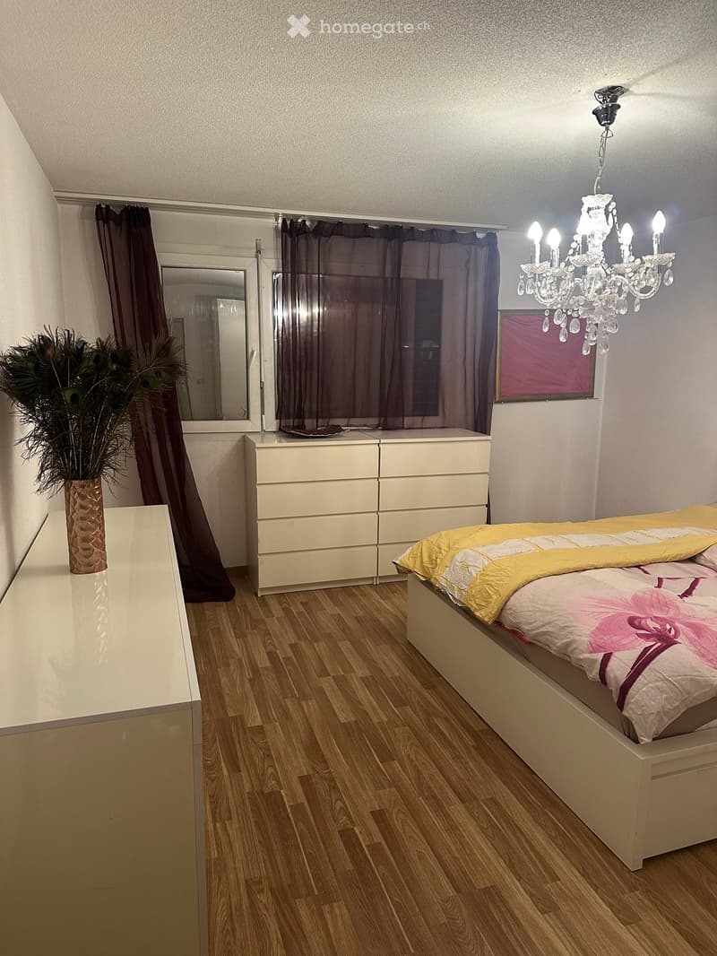 Modern bedroom with wooden flooring, a large bed with yellow and white bedding, a nightstand with a vase and plant, a mirror, and a chandelier.