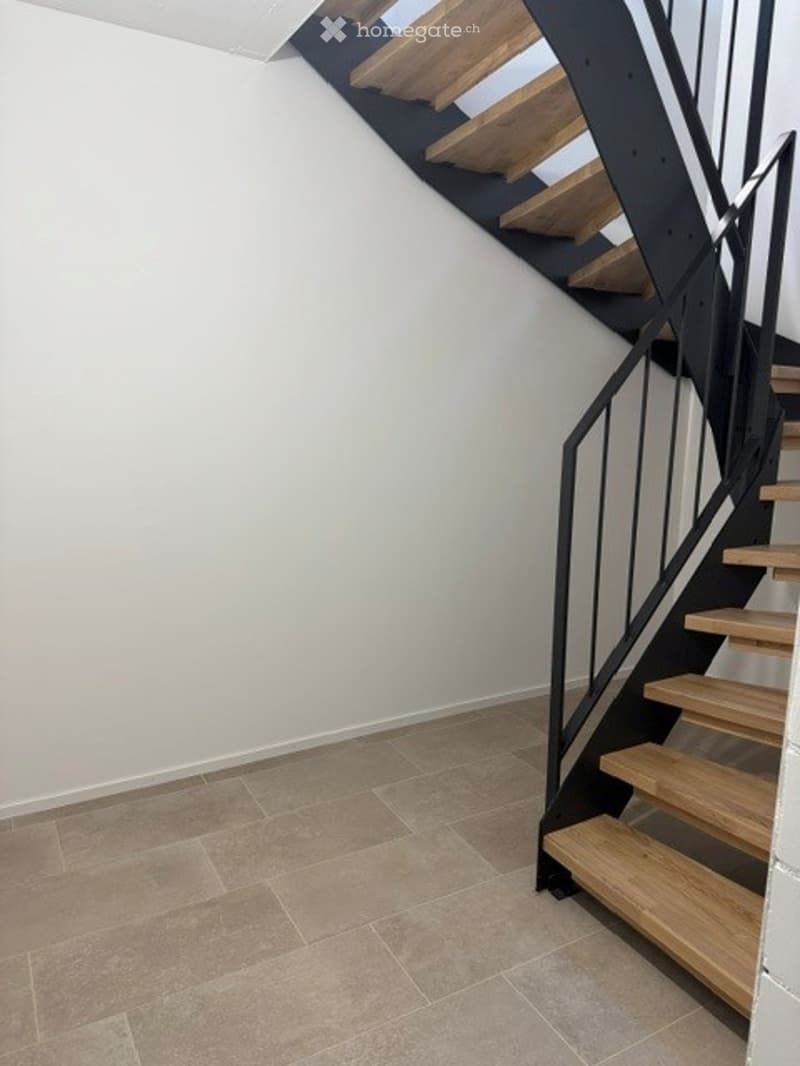 Modern staircase with wooden steps and black metal railing, beige tiled floor, white walls.