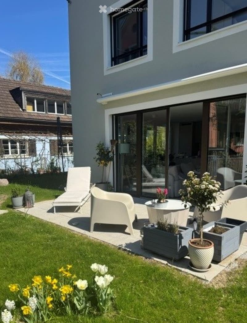 Well-maintained garden with a patio area, white lounge chairs, a small table, potted plants, and a view of the house with large sliding glass doors.