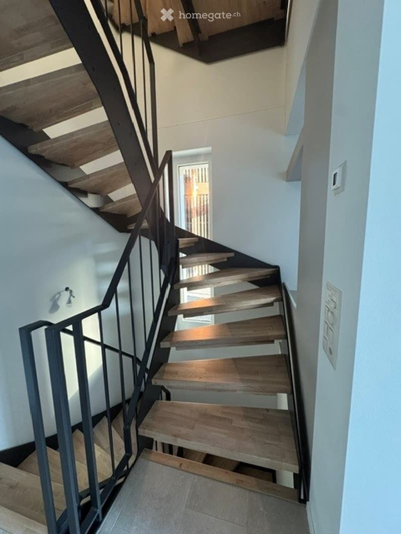 Modern staircase with wooden steps and metal railing, leading to an open area with natural light.