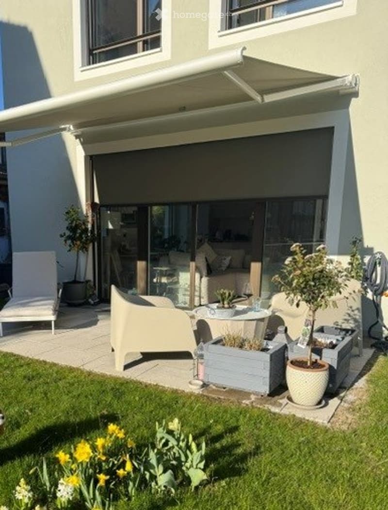 A modern patio area with a retractable awning, white lounge chairs, a small table, potted plants, and a well-maintained lawn with yellow flowers.