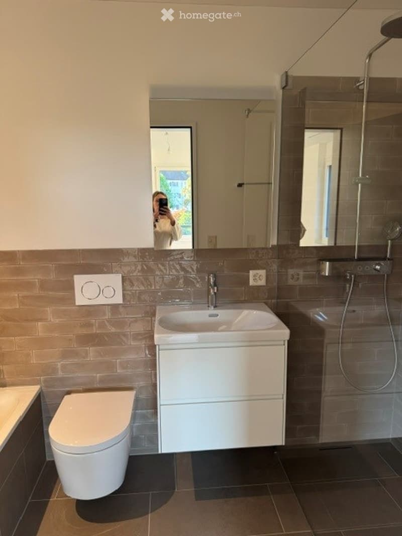 Modern bathroom with a white sink, toilet, and shower area. The walls are tiled in a combination of brown and beige, and there is a large mirror above the sink.