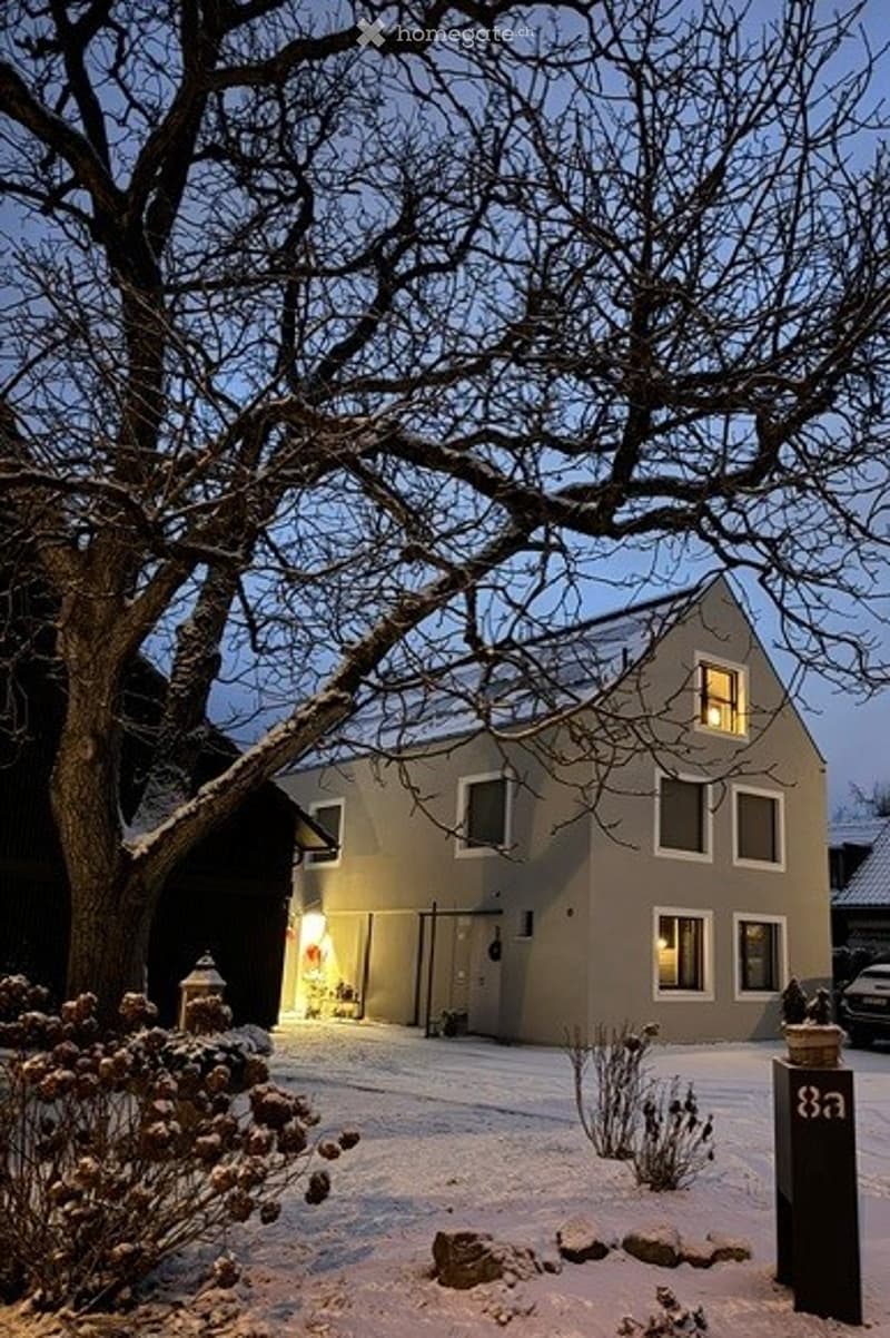 Two-story house with beige siding and white-framed windows, illuminated by warm interior lighting, surrounded by snow-covered ground and bare trees.