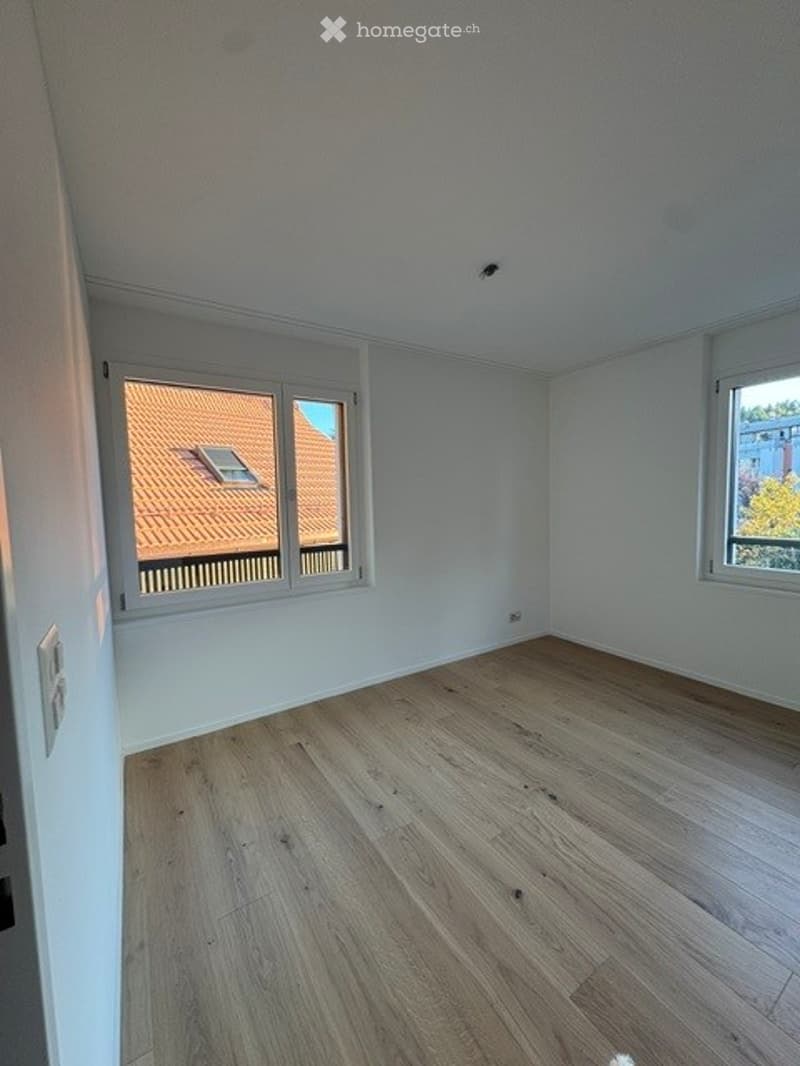 Empty room with light wooden flooring, white walls, and large windows showing a view of a neighboring roof and greenery.