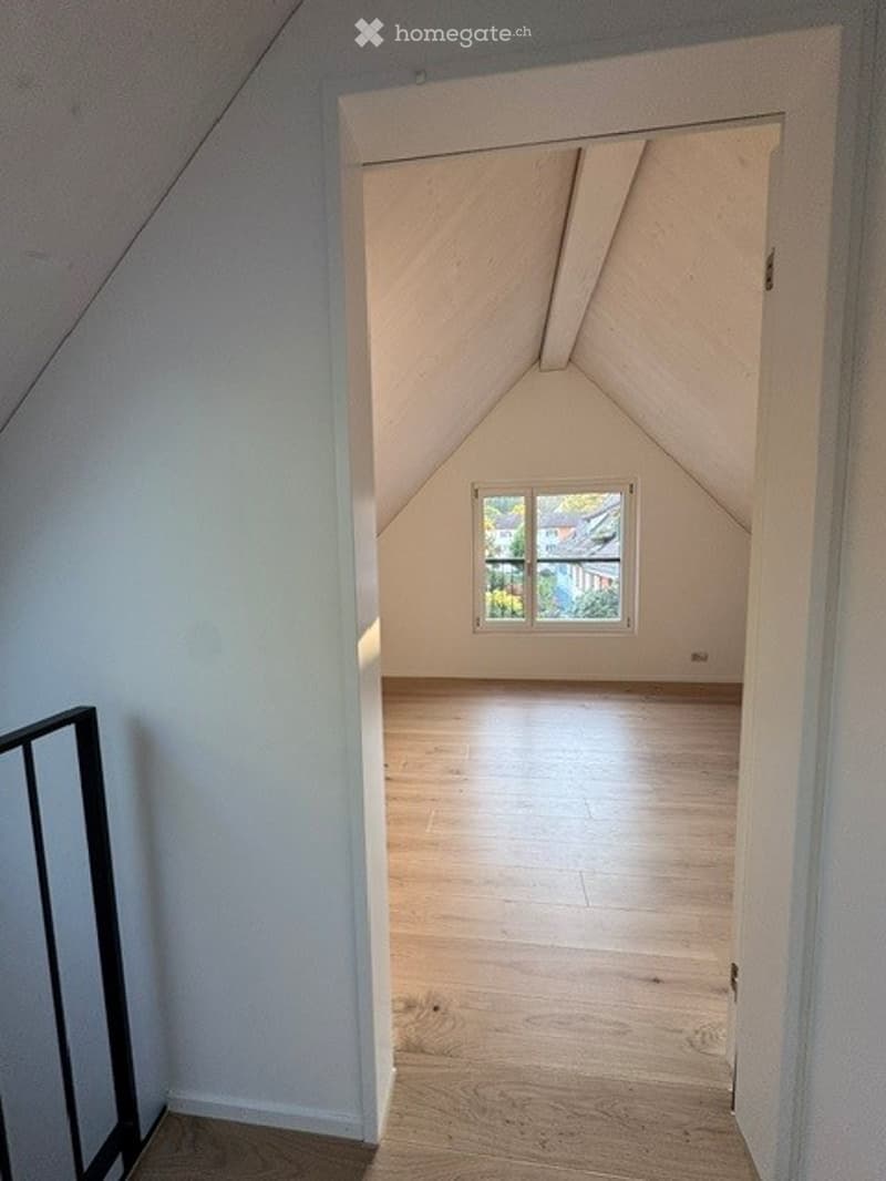 Empty attic room with wooden flooring, white walls, and a single window letting in natural light. The sloped ceiling features exposed wooden beams.