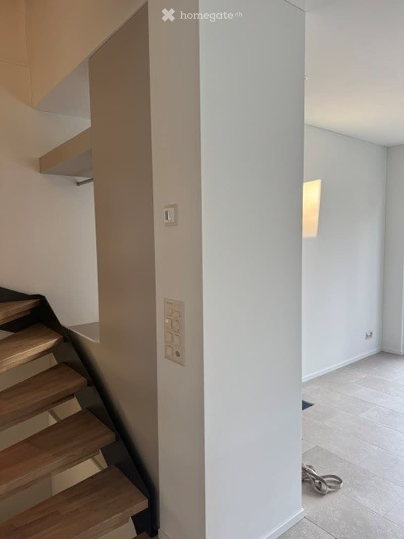 A modern hallway with a wooden staircase, white walls, and a small shelf on the left side. The floor is tiled, and there is a light switch on the wall.