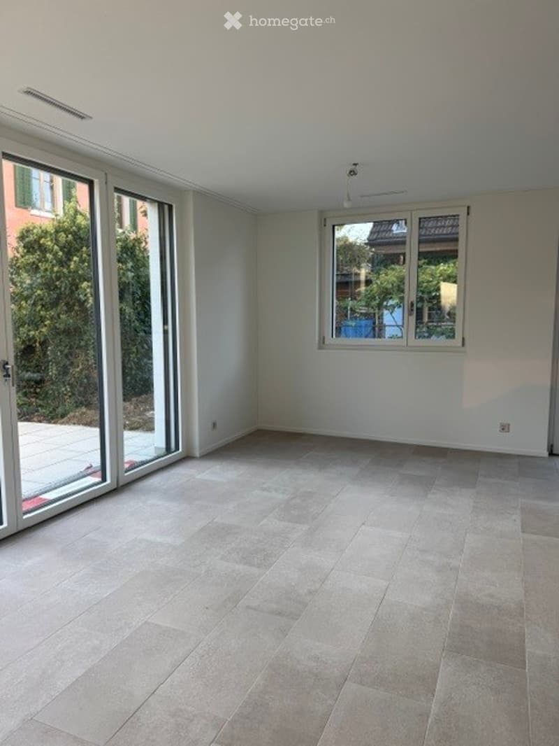 Empty room with large sliding glass doors, light grey tiled floor, white walls, and a window with a view of greenery.