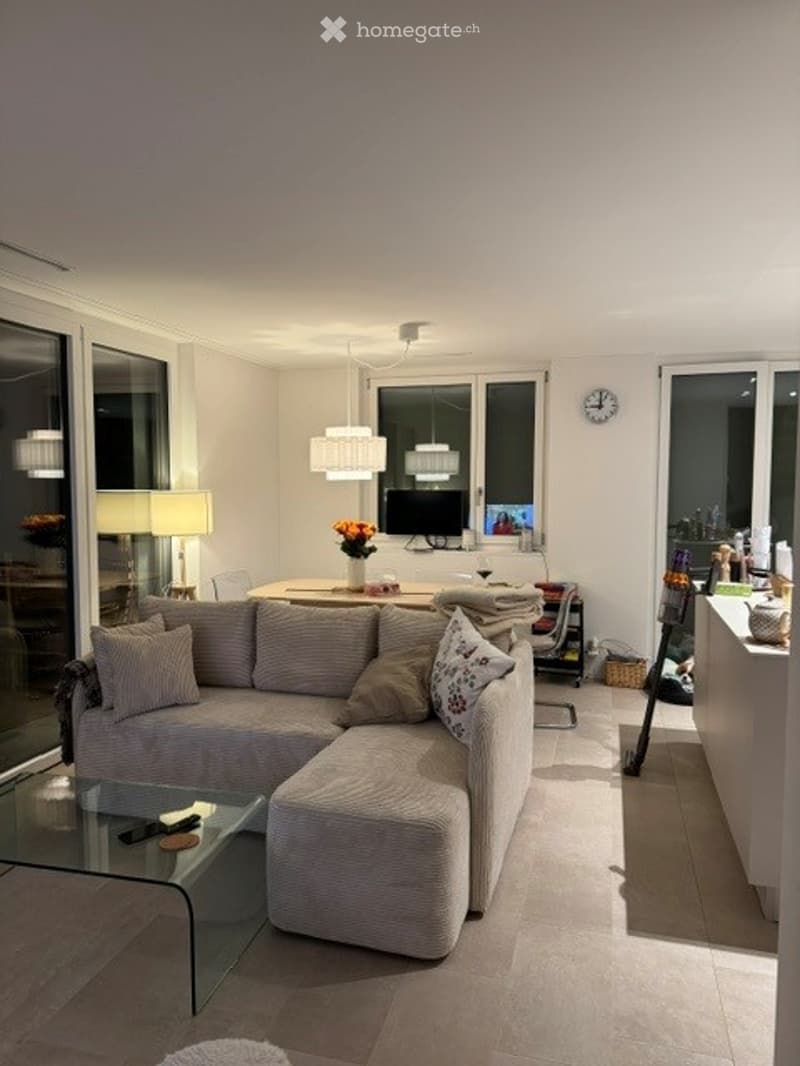 Modern living room with a grey sectional sofa, coffee table, dining table, chairs, lamps, and a television. The space features large windows and a sleek design.