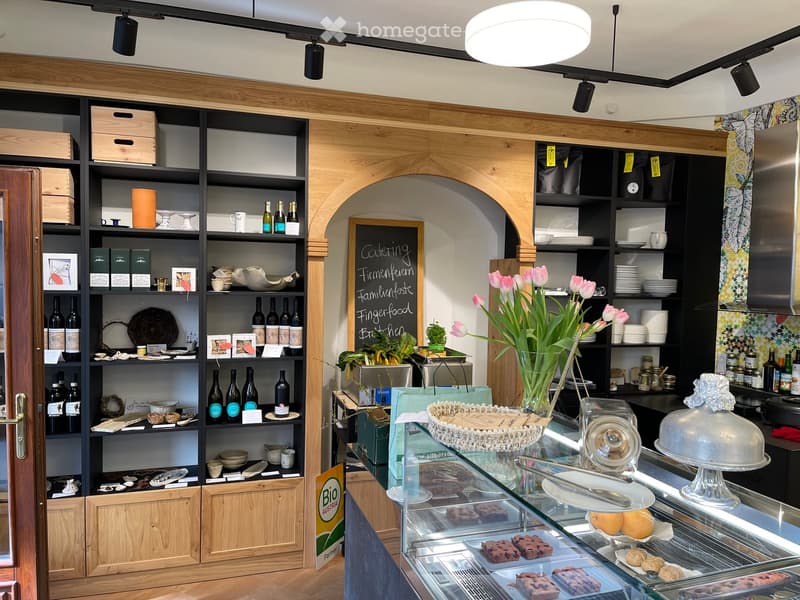 A well-organized shop interior with wooden shelves filled with wine bottles, decorative items, and plates. A display case in the foreground holds cakes and pastries, and a vase with pink flowers sits on the counter.