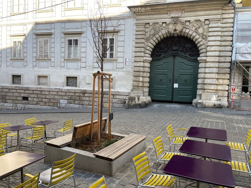 Outdoor seating area with tables and chairs arranged around a small tree, adjacent to a historic building with a large green door.
