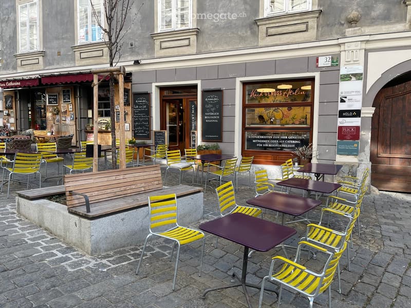 Outdoor seating area with tables, chairs, and a bench in front of a building with storefronts.