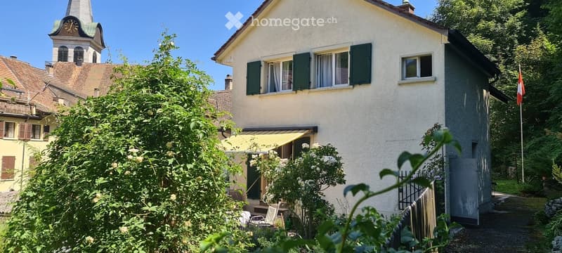 A two-story house with white walls, green shutters, and a yellow awning over the entrance. The house is surrounded by lush greenery and features a small garden area with outdoor furniture.