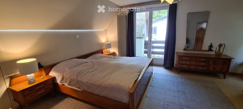 Cozy bedroom with a wooden bed, nightstands with lamps, a dresser with a mirror, and a rug on the floor. Large window with curtains provides natural light.