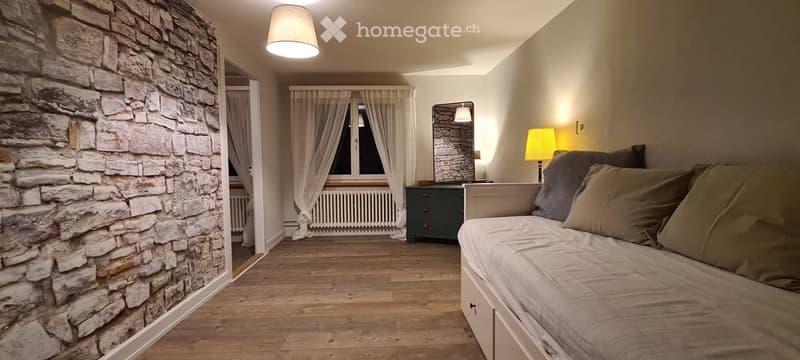 Cozy bedroom with a daybed, wooden flooring, stone accent wall, and soft lighting. Features include a mirror, side table, and a radiator beneath the window.