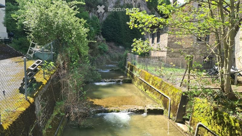 A narrow, mossy canal flows through a lush, green area with trees and a stone building in the background. The canal has small waterfalls and is bordered by a metal fence and wooden planks.