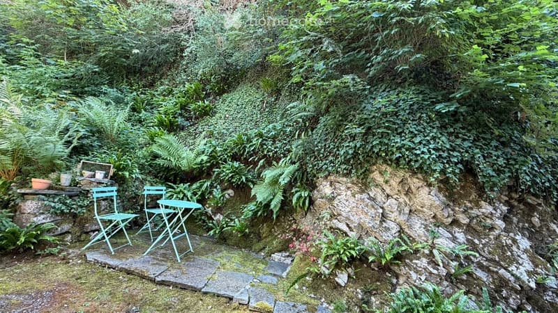A small, lush garden area with a stone pathway, a small table and chairs, surrounded by dense greenery and ferns.
