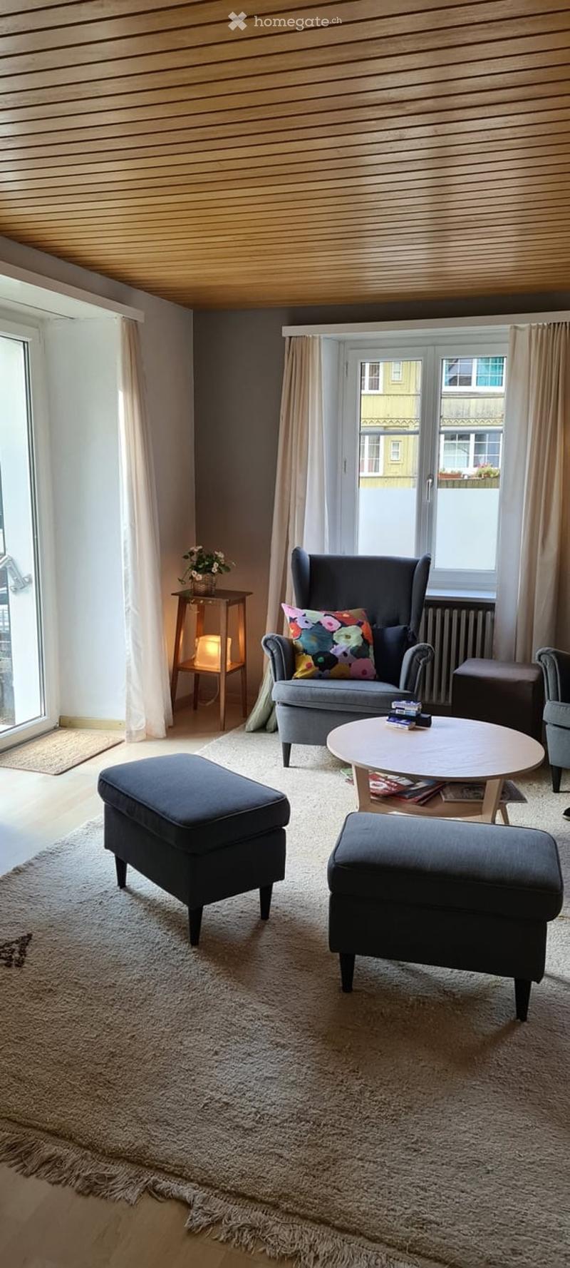 Modern living room with large windows, grey sofa, coffee table, ottomans, side table with a lamp, and a colorful throw pillow.