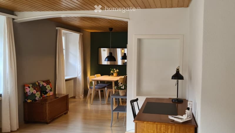 Open-plan living and dining area with wooden flooring, a sofa with colorful cushions, a dining table with chairs, a desk with a lamp, and a mirror on the wall.