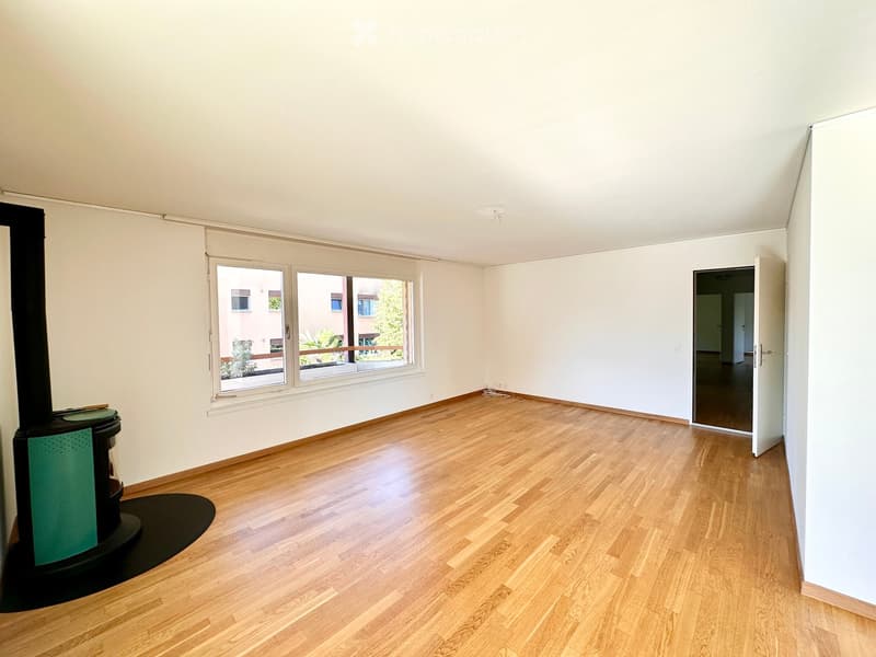 Empty room with light-colored walls, wooden flooring, and a fireplace on the left side. There are large windows with a view of the outside and an open doorway leading to another room.