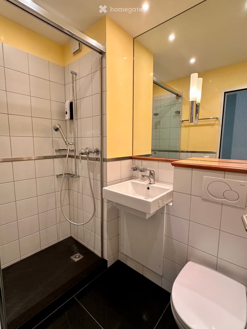 Modern bathroom with white tiles, a shower stall, a sink with a modern faucet, a toilet, and a large mirror with built-in lighting.