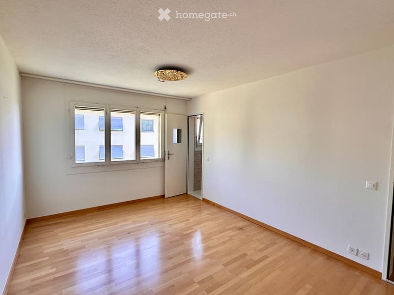 Empty room with wooden flooring, white walls, and large windows with blinds. A door leads to another room, and a ceiling light fixture is visible.