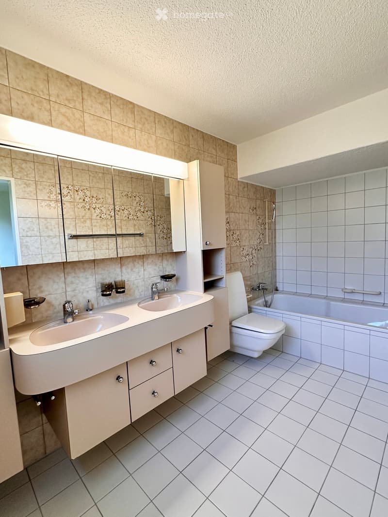 Modern bathroom with white tiled walls and floor, double sink vanity with mirrored cabinet, toilet, bathtub, and built-in storage.