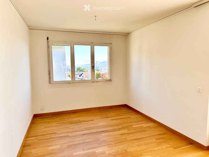 Empty room with wooden flooring and white walls, featuring large windows with a view of the city and mountains.