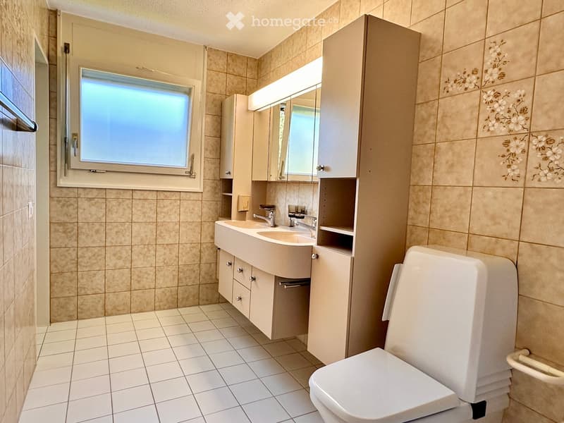 Modern bathroom with beige tiles, double sink, mirror, and toilet. Features a window and built-in cabinets.