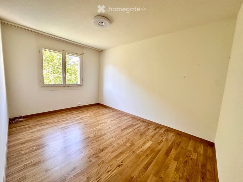 Empty room with wooden flooring, white walls, and a window with white frames. The room has a ceiling light fixture and appears to be a blank canvas for furnishing.