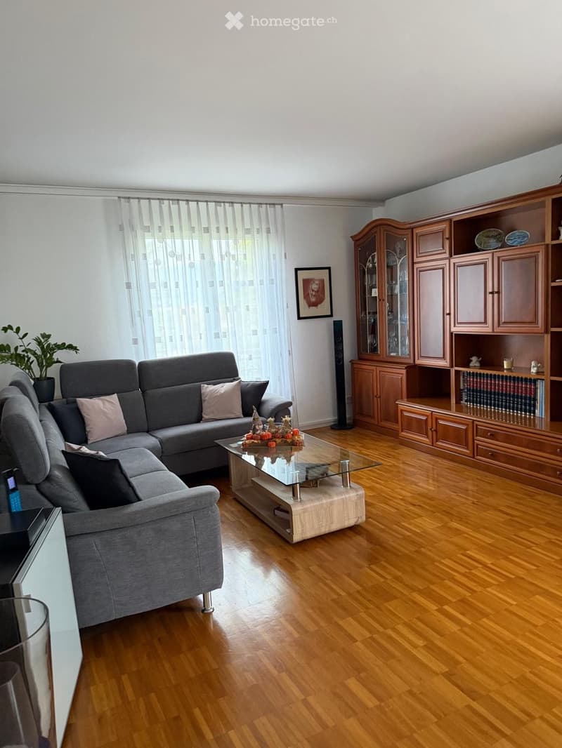 Modern living room with wooden flooring, grey sectional sofa, glass coffee table, wooden bookshelf, and decorative items.