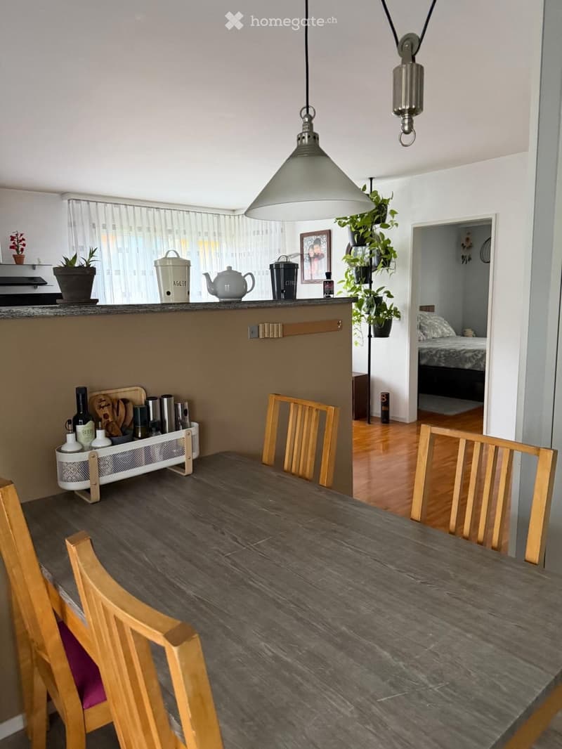 Dining area with a wooden table and chairs, a small kitchen counter with utensils, and a view into a bedroom with a bed.
