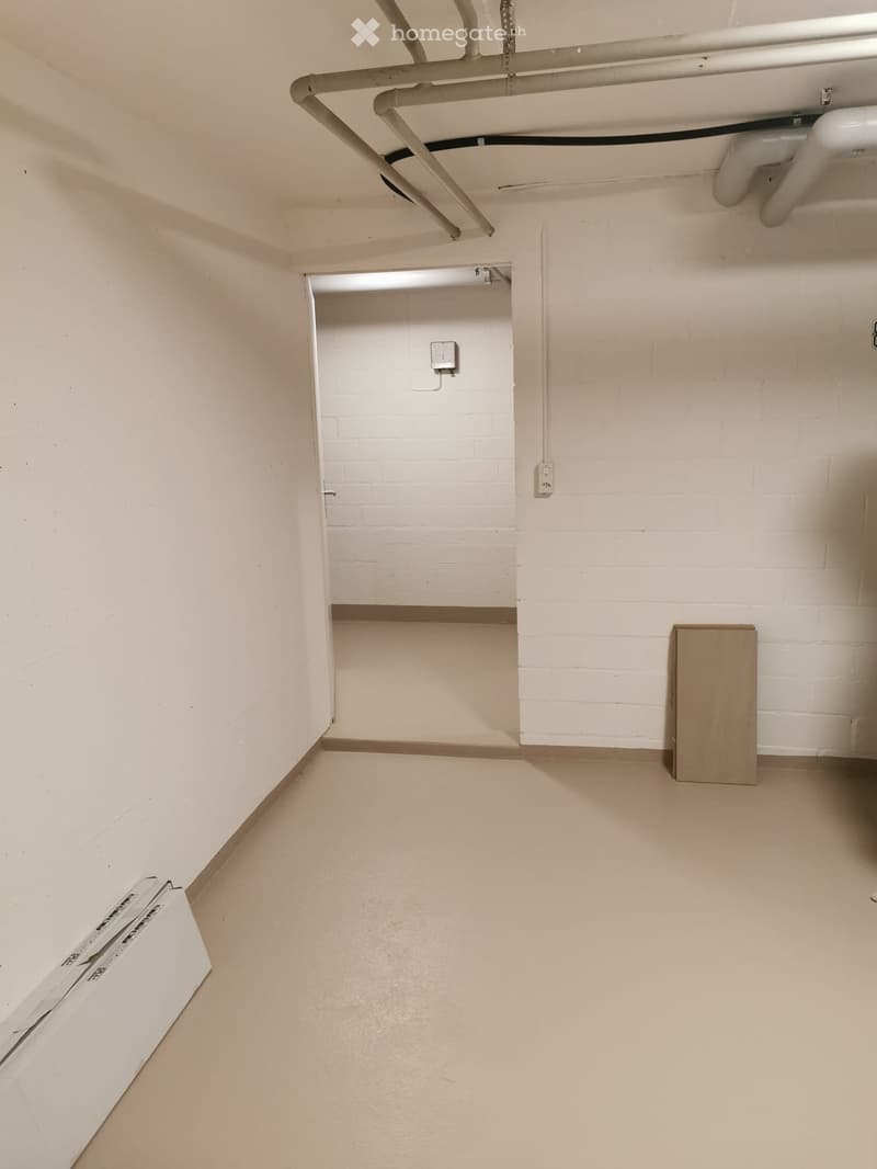 Empty room with beige walls, a concrete floor, and a small wooden cabinet in the corner. Exposed pipes and electrical outlets are visible on the walls.