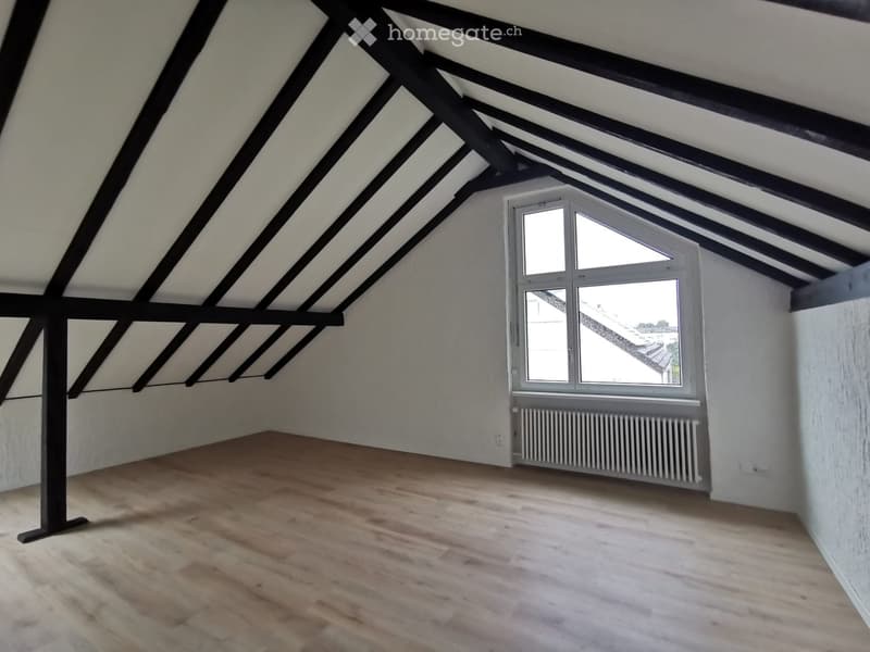 Empty attic room with wooden flooring, exposed wooden beams, and a single window with white frame and radiator below.