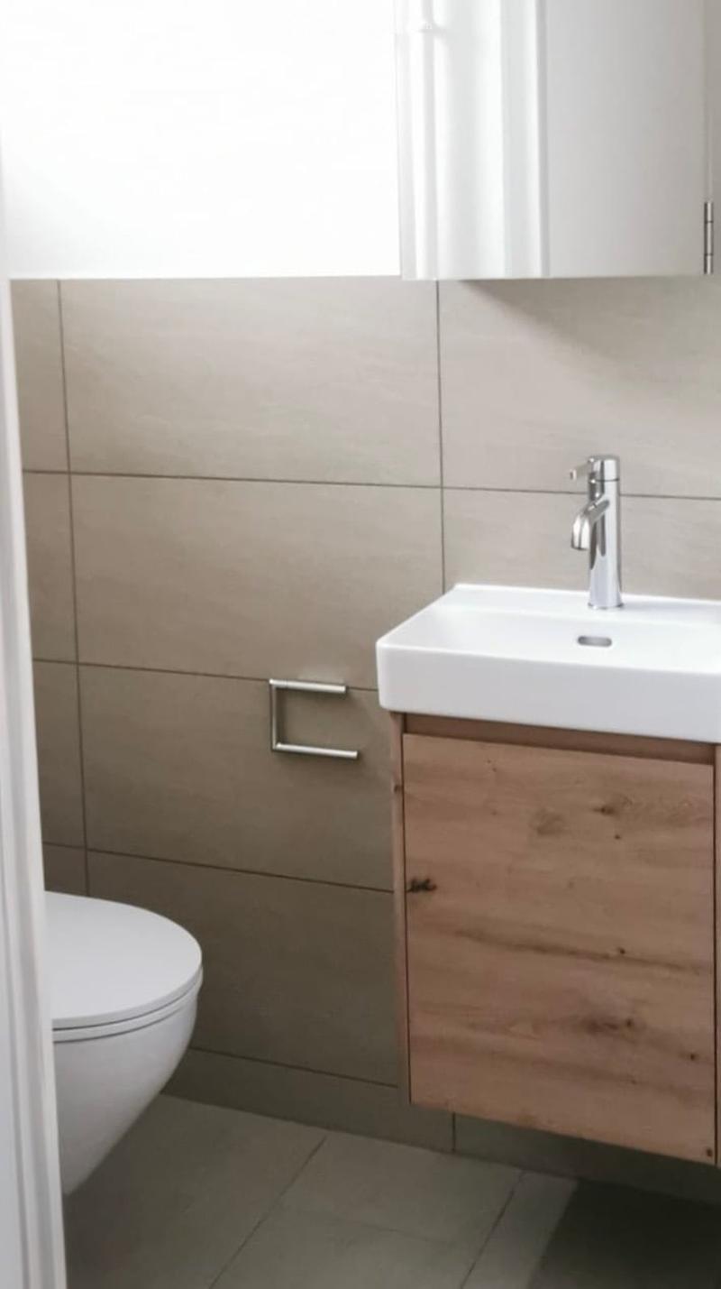 Modern bathroom with a white toilet, a white sink with a chrome faucet, and a wooden cabinet. The walls are tiled in a neutral color scheme, and the floor is made of dark tiles.