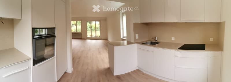 Open-plan kitchen and living area with white cabinets, light wood flooring, and large windows. The kitchen features a sink, stove, and built-in oven, while the living area has a spacious layout with natural light.