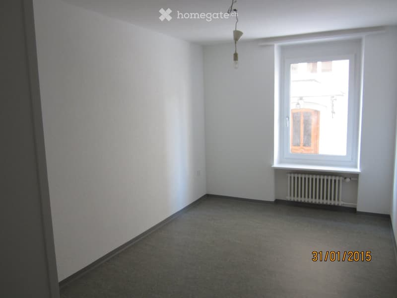 Unfurnished Room. Empty room with white walls, grey carpeted floor, single window with white frame, radiator below window, and a hanging light fixture.
