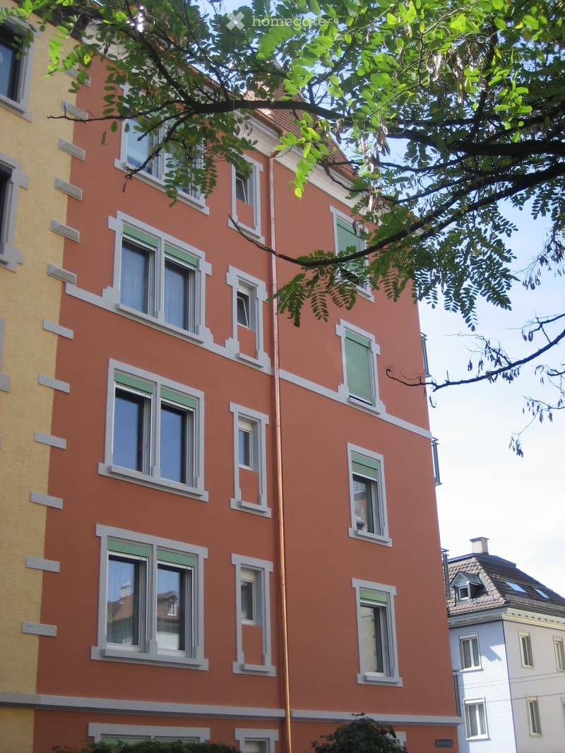 Building. A multi-story residential building with orange and yellow facades, white window frames, and green shutters. The building has multiple windows and is partially shaded by tree branches.