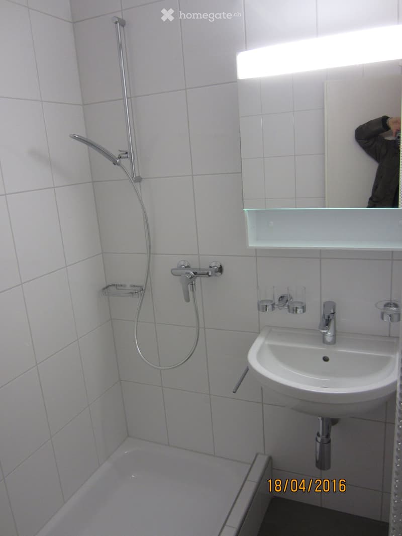 Bathroom. Modern bathroom with white tiled walls, a sink with a chrome faucet, a shower with a handheld sprayer, a glass shelf above the sink, and a mirror.