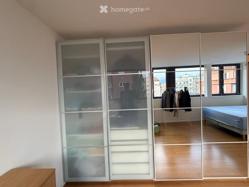 Modern bedroom with wooden flooring, large mirrored sliding wardrobe, bed, and clothing rack with jackets.
