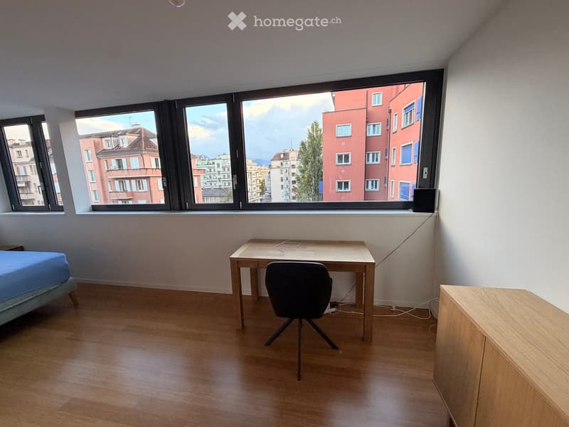 A furnished room with a bed, desk, chair, and cabinet, featuring large windows with a cityscape view.