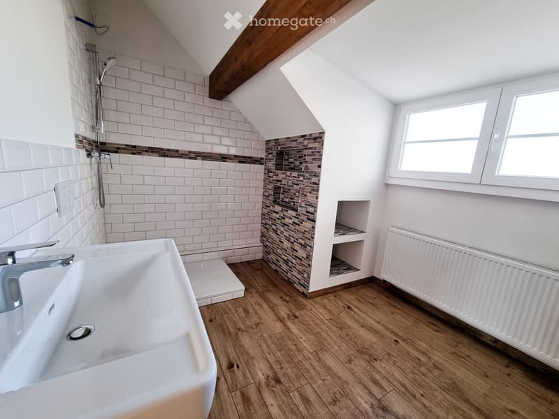 Modern bathroom with white tiles, wooden flooring, a large sink, a shower area, and a radiator. The room features a skylight window and built-in shelving.
