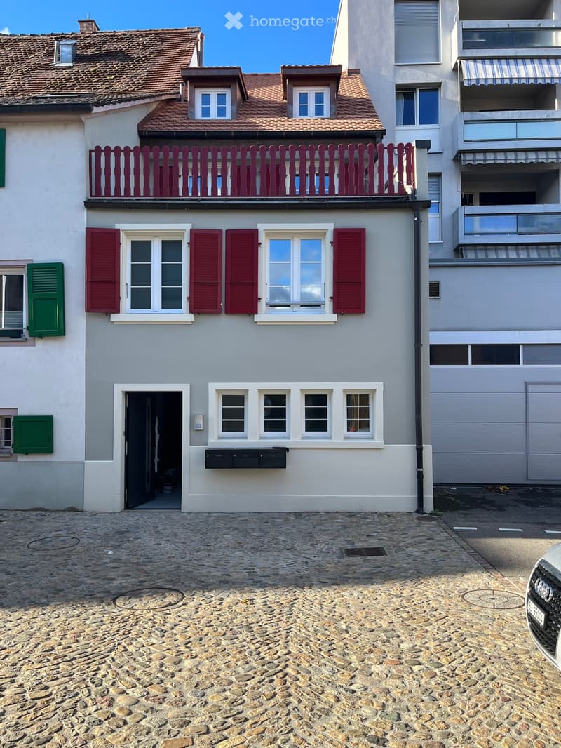 Two-story house with red shutters, white windows, and a red balcony railing. The ground floor has a door and several windows, while the upper floor features additional windows and a small balcony. The house is situated on a cobblestone street.