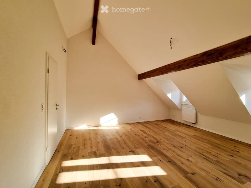 Empty attic room with wooden flooring, slanted ceilings, and exposed wooden beams. Natural light streams in from a skylight window.