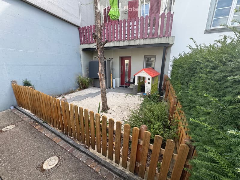 Small fenced garden area with a tree, a playhouse, and a chair near the entrance of a building.