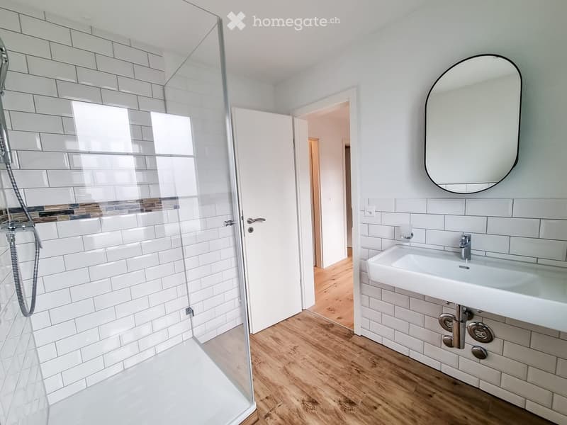 Modern bathroom with white tiled walls, a glass-enclosed shower, a white sink with a modern faucet, and a large round mirror above the sink.