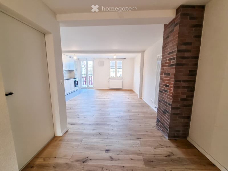 Empty room with wooden flooring, white walls, and a brick pillar. The room is connected to a kitchen area with white cabinets and a window.