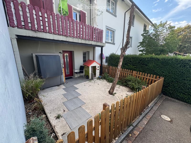 Small garden area with gravel ground, wooden fence, small playhouse, and a chair near the entrance of a building.